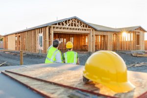 Two construction workers in high-visibility vests and yellow hard hats stand in front of a partially framed wooden house at sunset.